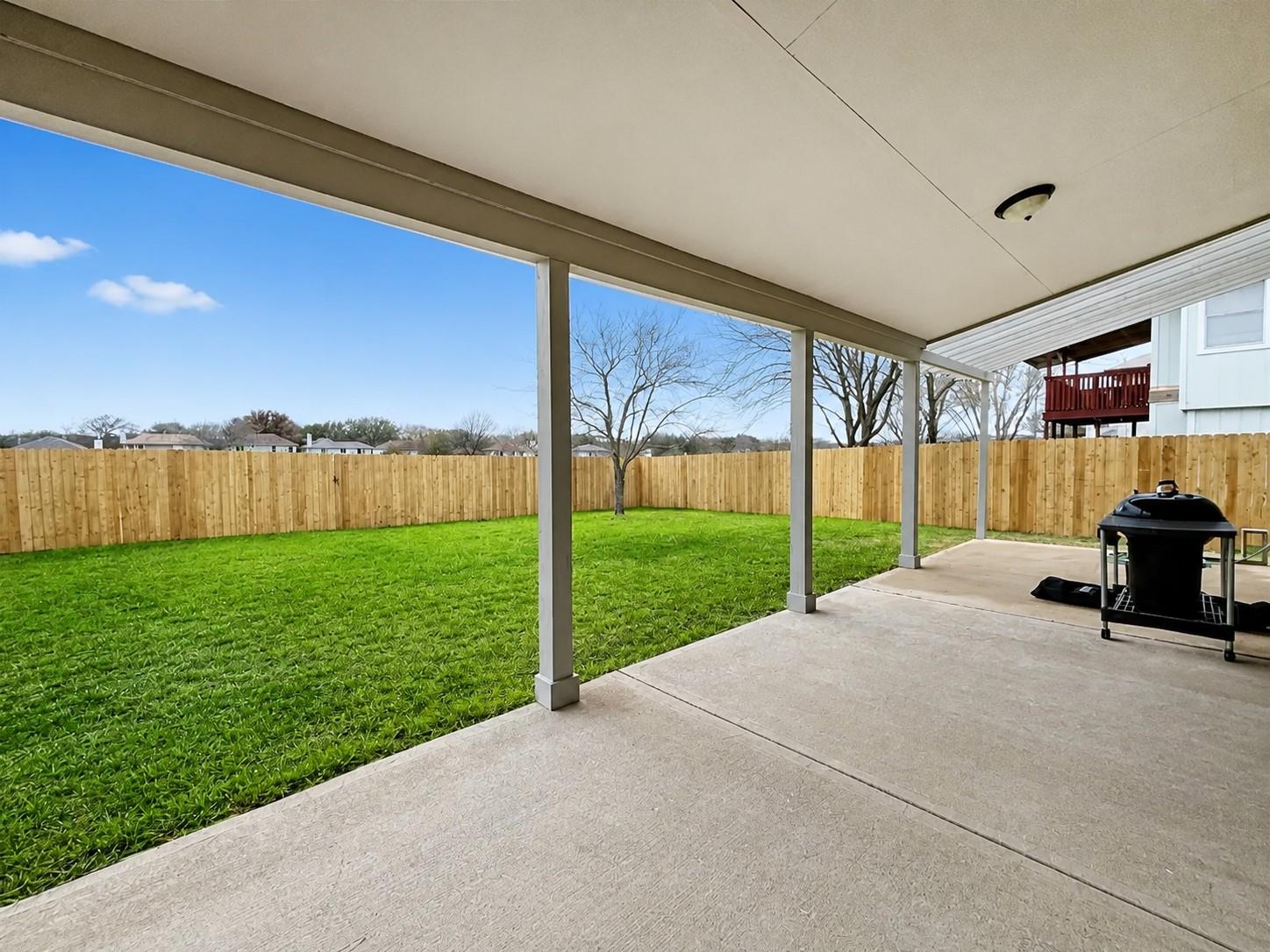 Covered concrete patio with overhead lighting, overlooking a green lawn, wood privacy fencing, and mature trees