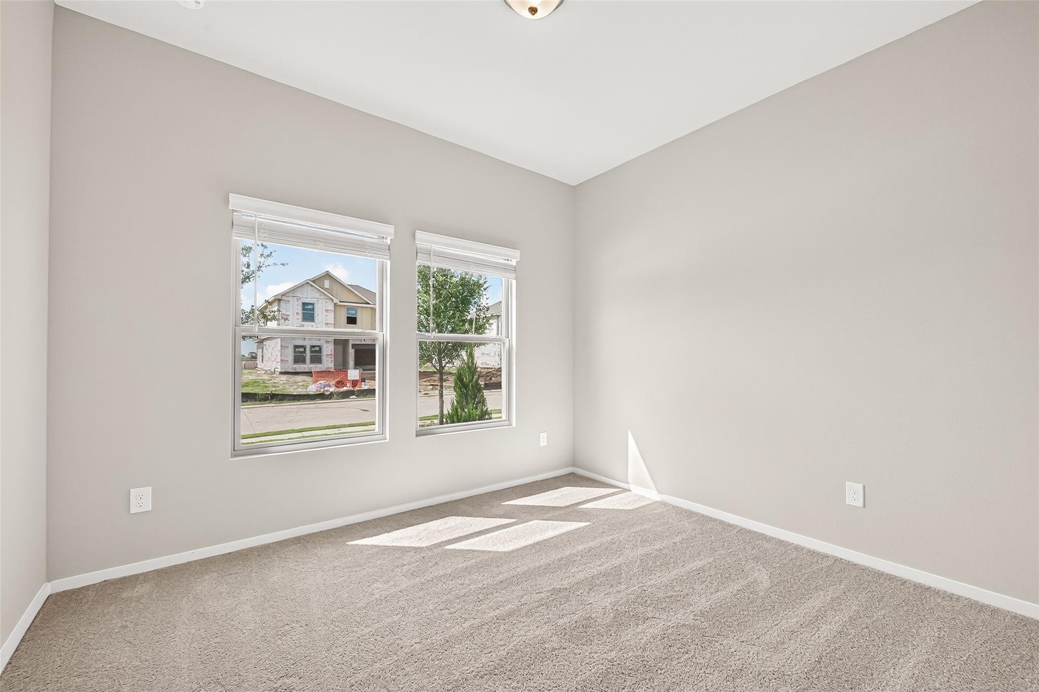 Bright and airy room with neutral walls and carpet, featuring two large windows offering plenty of natural light and a view of the neighborhood.