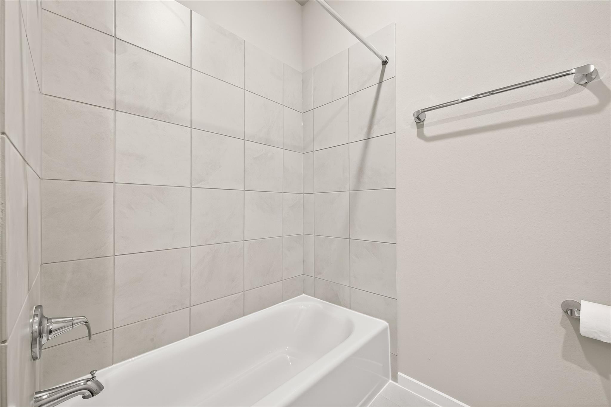 This photo shows a clean, modern bathroom featuring a white bathtub with a tiled shower area in neutral tones. It includes a wall-mounted towel rack and a toilet paper holder for convenience.
