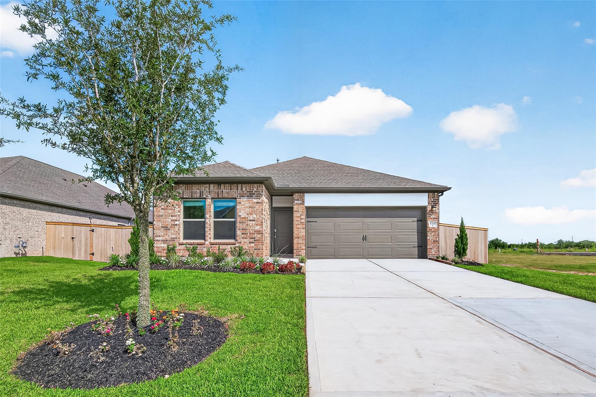 The house showcases a modern, single-story brick home with a well-maintained front yard, a young tree, and a spacious driveway leading to a two-car garage. The property features landscaped flower beds and is set in a quiet neighborhood.