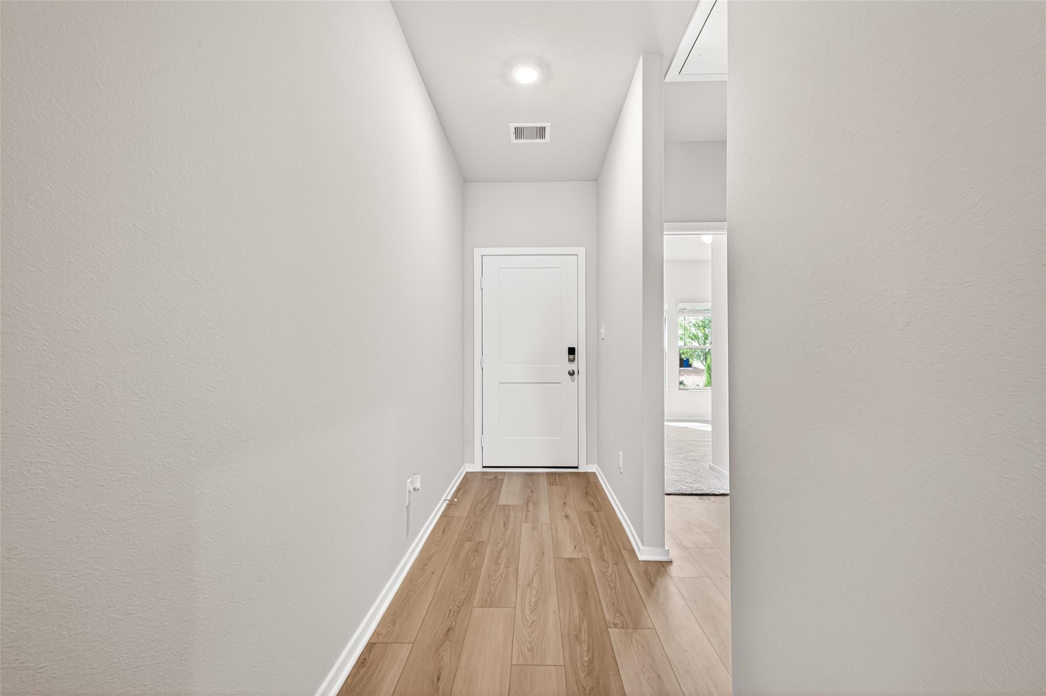 Bright, modern hallway with light wood flooring and neutral walls, leading to a white door. A side doorway opens to a room with a window view.