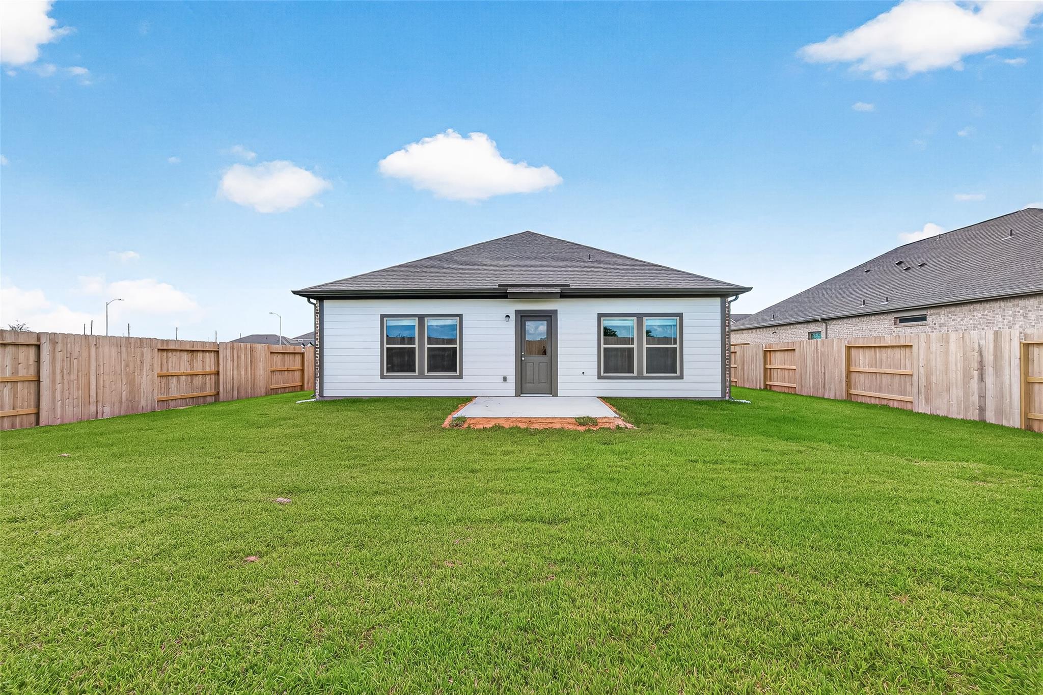This photo shows the back of a modern single-story home with a spacious fenced yard and a small patio. The exterior is light-colored with a dark roof, and there are multiple windows for natural light.