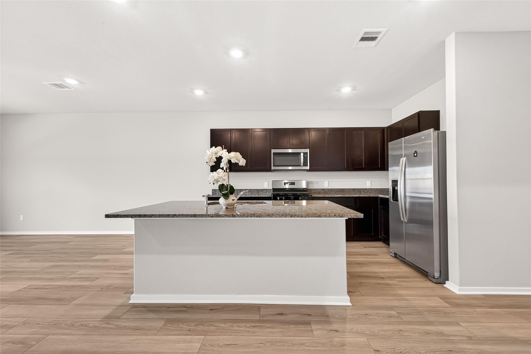 Modern kitchen with an open layout, featuring dark wood cabinets, stainless steel appliances, and a large island with granite countertops. The space is bright with recessed lighting and light wood flooring.