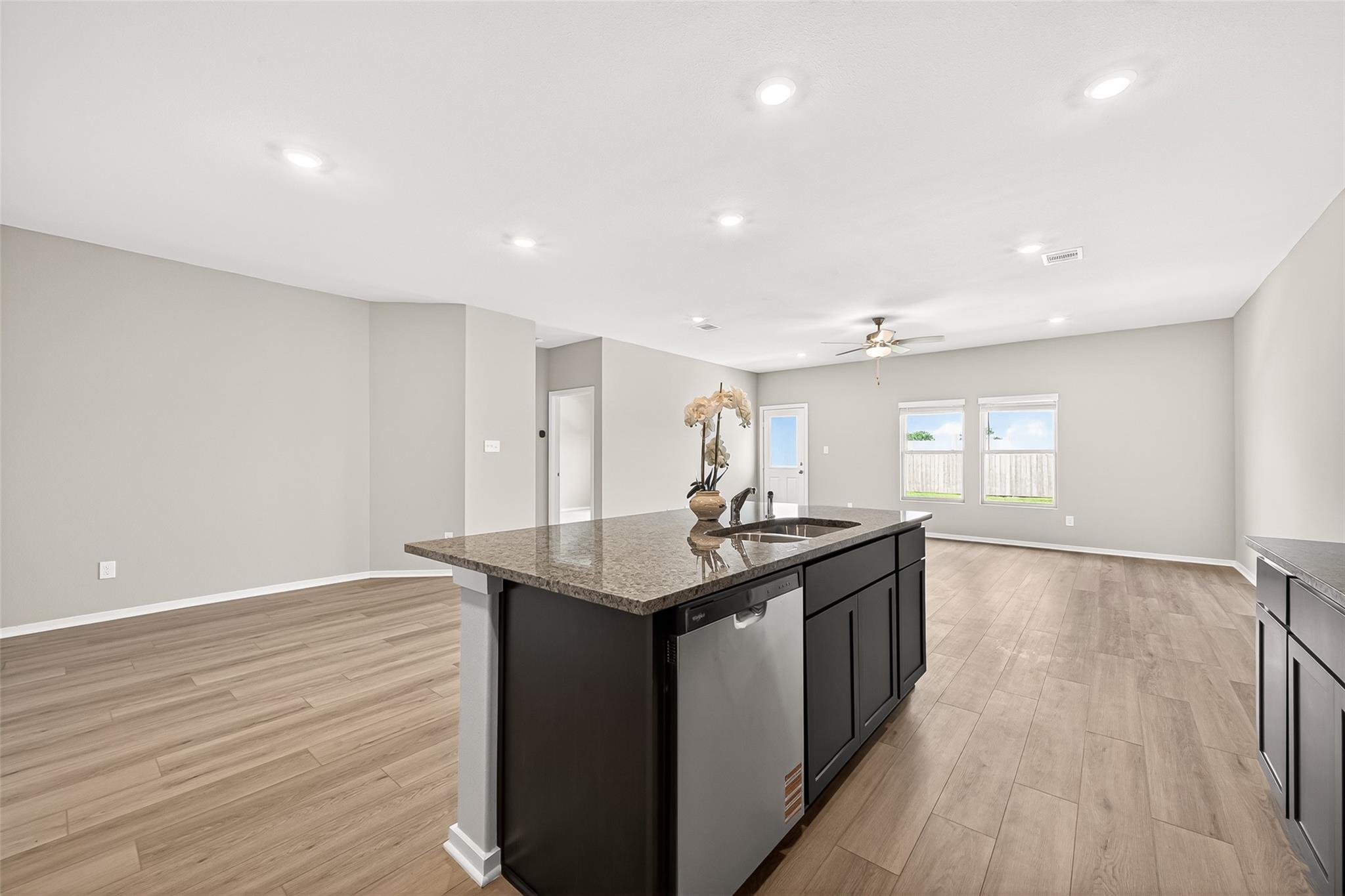 This open-concept kitchen and living area features modern recessed lighting, sleek countertops, and an island with a built-in sink. The room is spacious with light wood flooring, neutral walls, and abundant natural light from large windows.