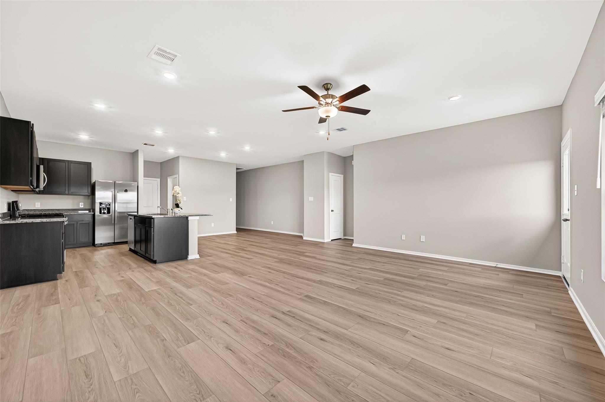 A spacious, open-concept living area with modern wood flooring and neutral walls. The kitchen features dark cabinets, stainless steel appliances, and a central island with a sink. A ceiling fan is centrally located, enhancing the bright, airy feel.