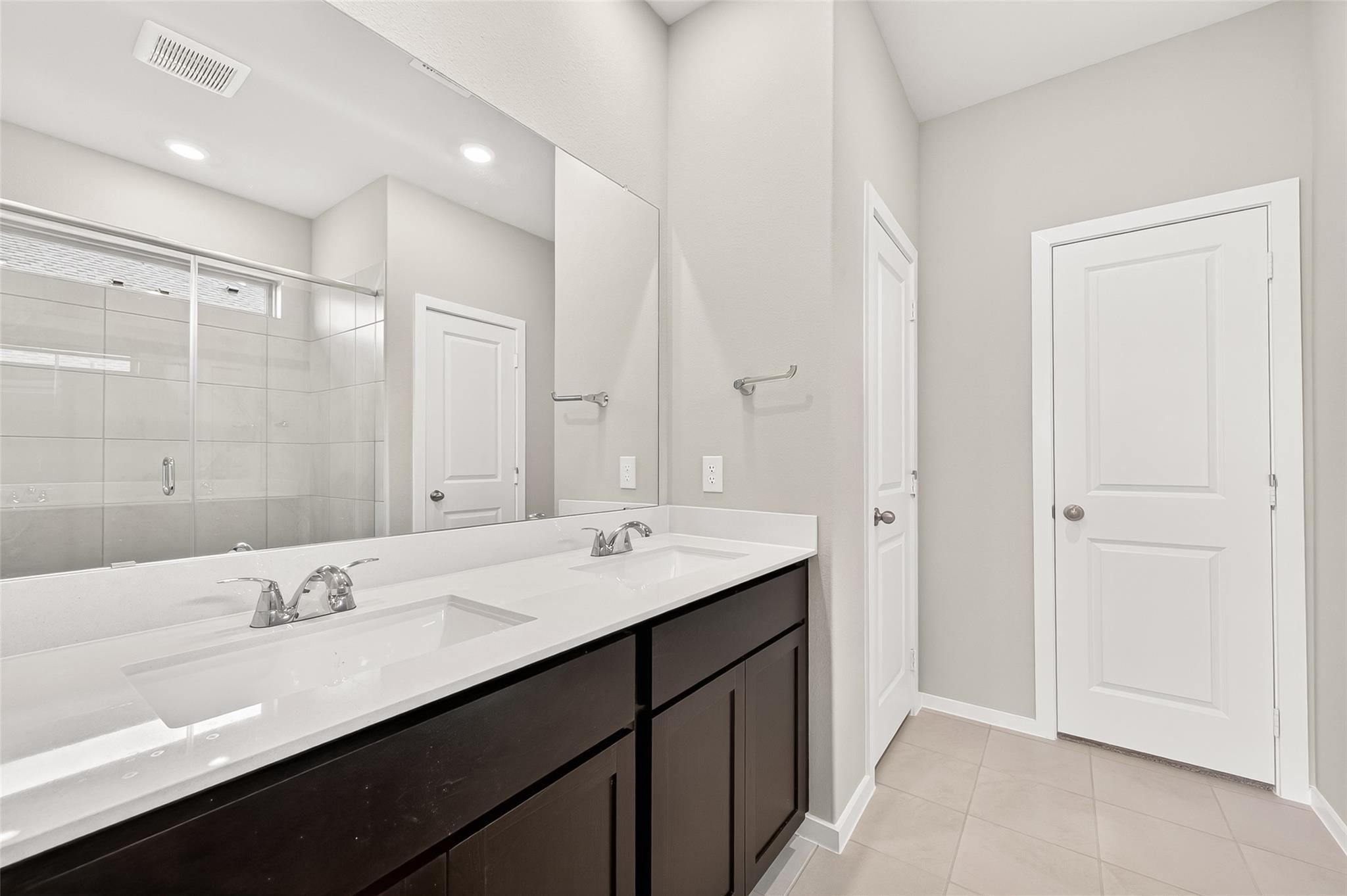 This photo features a modern bathroom with a double sink vanity, sleek black cabinetry, and a large mirror. The room is well-lit with recessed lighting and features a spacious glass-enclosed shower. Neutral tones create a clean, contemporary look.