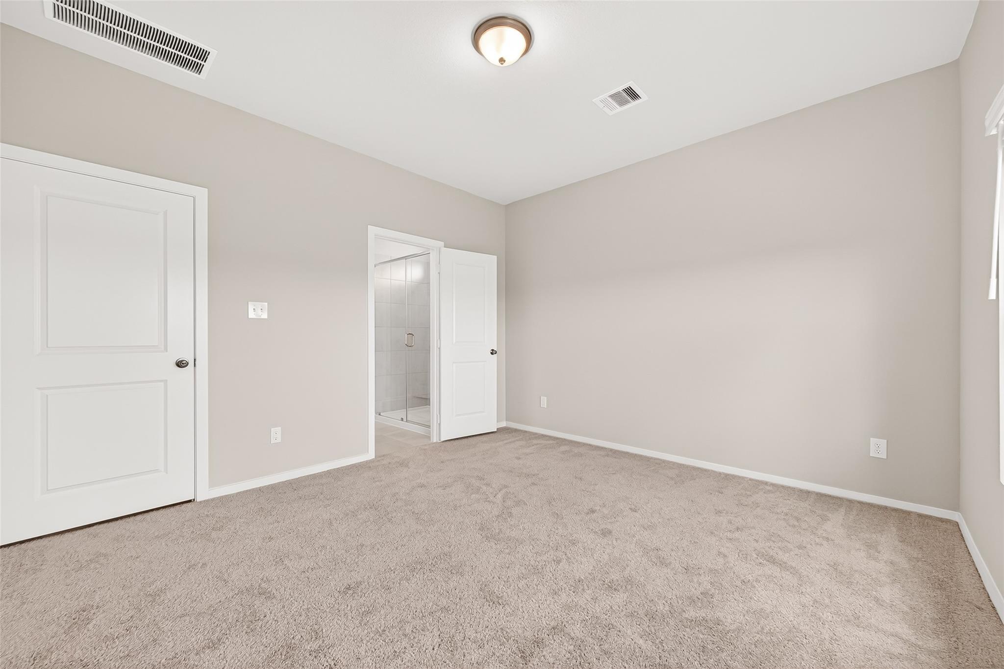 This photo shows a spacious, neutral-toned bedroom with carpeted flooring, a ceiling light fixture, and two doors, one leading to a tiled bathroom. The room is well-lit with natural light.