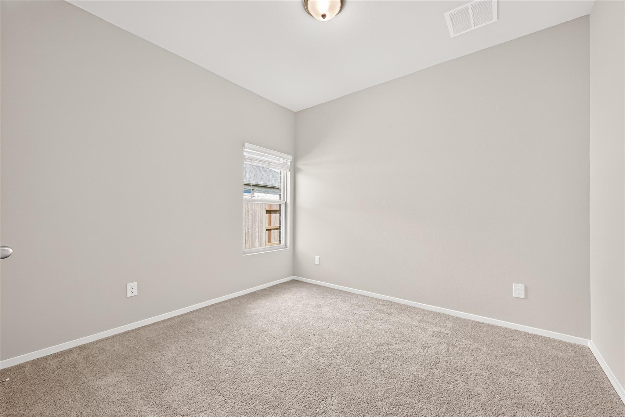 This photo showcases a small, bright room with neutral-toned walls and carpet flooring. It features a single window allowing natural light and a ceiling light fixture, ideal for a bedroom or office space.