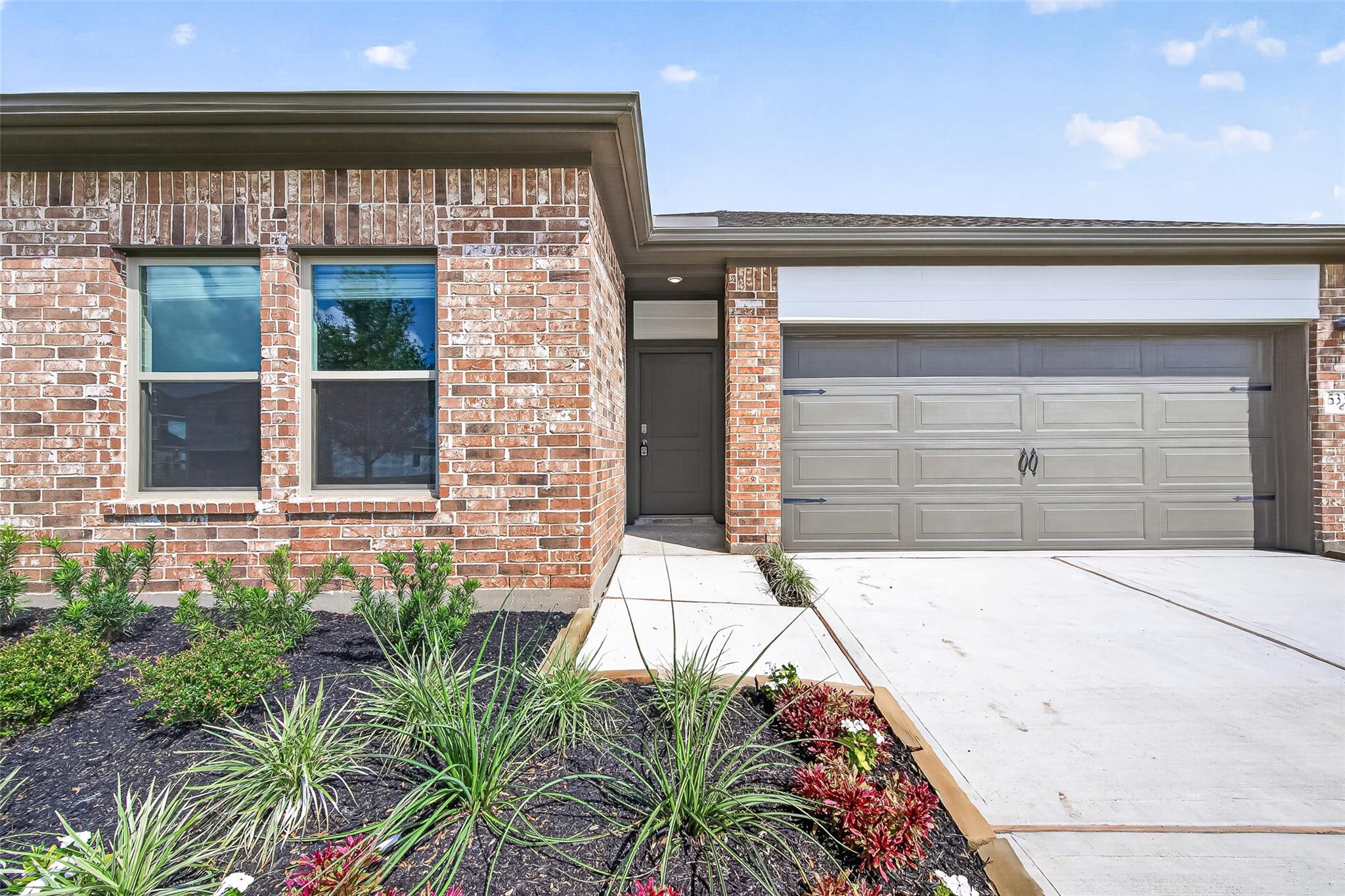 This photo showcases a brick home with a modern facade, featuring a two-car garage and a well-maintained garden with various plants. The entrance is inviting, with a paved walkway leading to the front door.