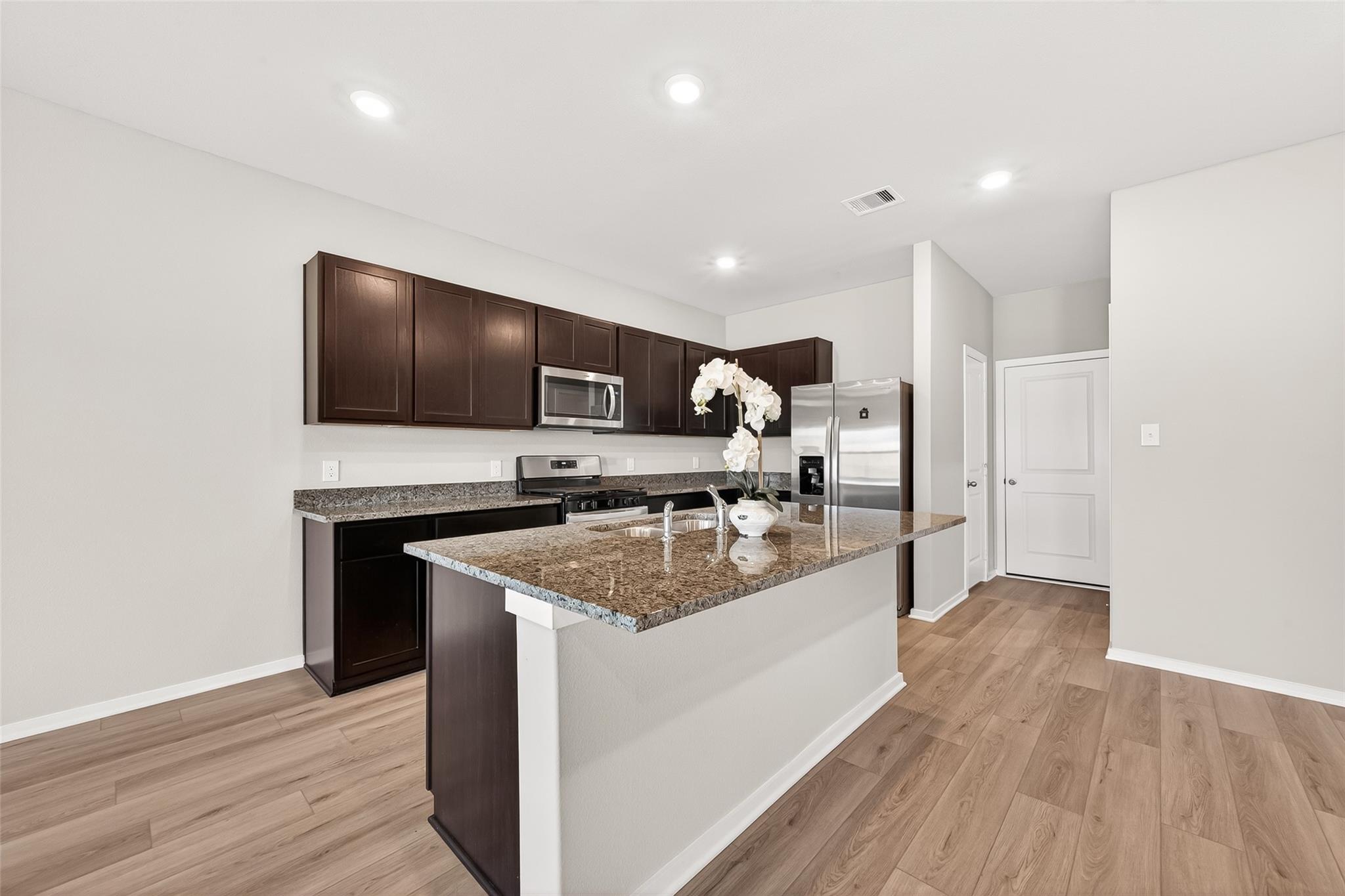 This kitchen features modern stainless steel appliances, dark wood cabinetry, and a granite-topped island. The open layout and light wood floors create a bright, inviting space.