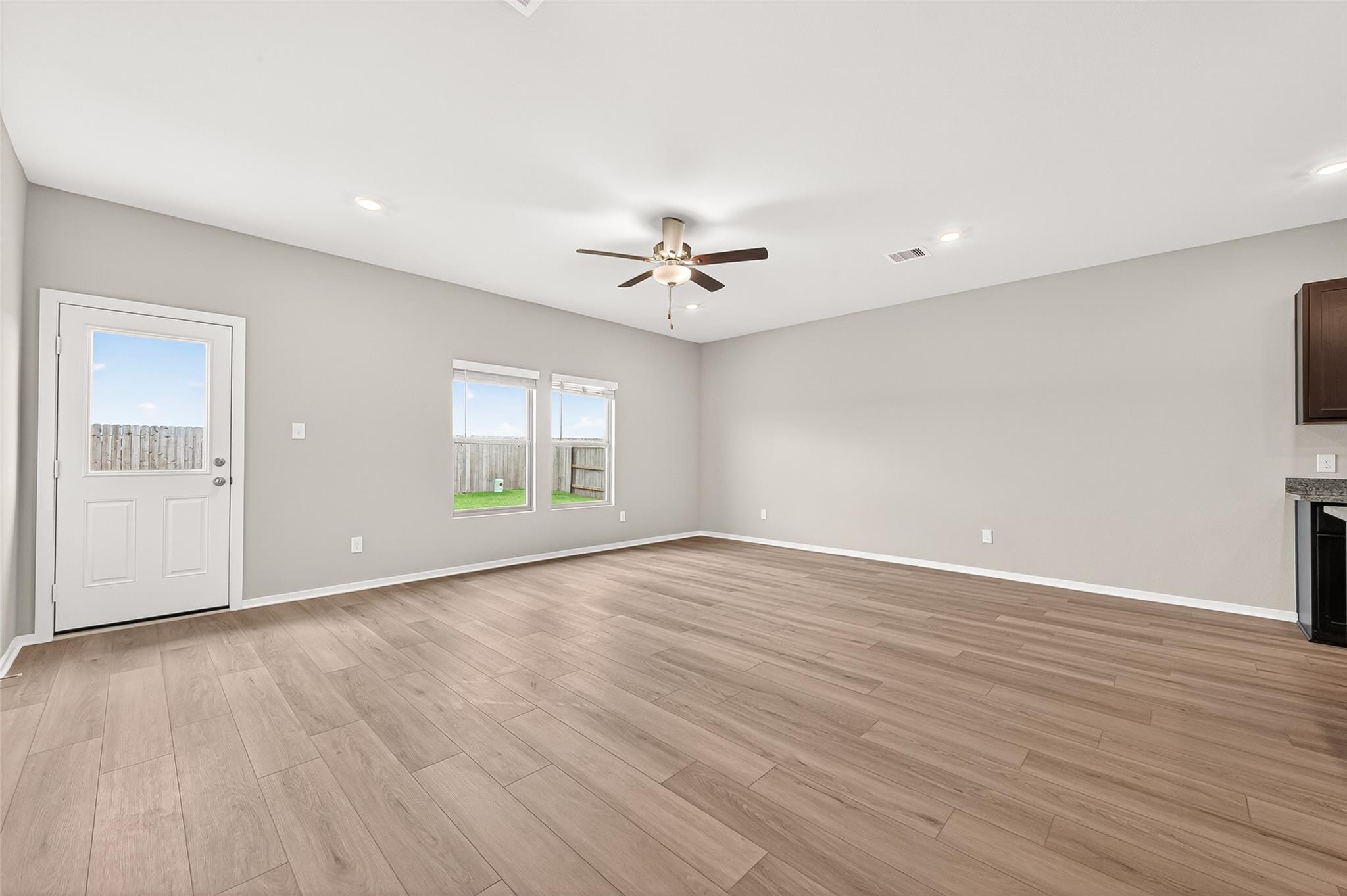 A spacious living area features light wood flooring, neutral walls, a ceiling fan, and ample natural light from large windows. The room also includes a door leading to the backyard, creating a bright and inviting space.