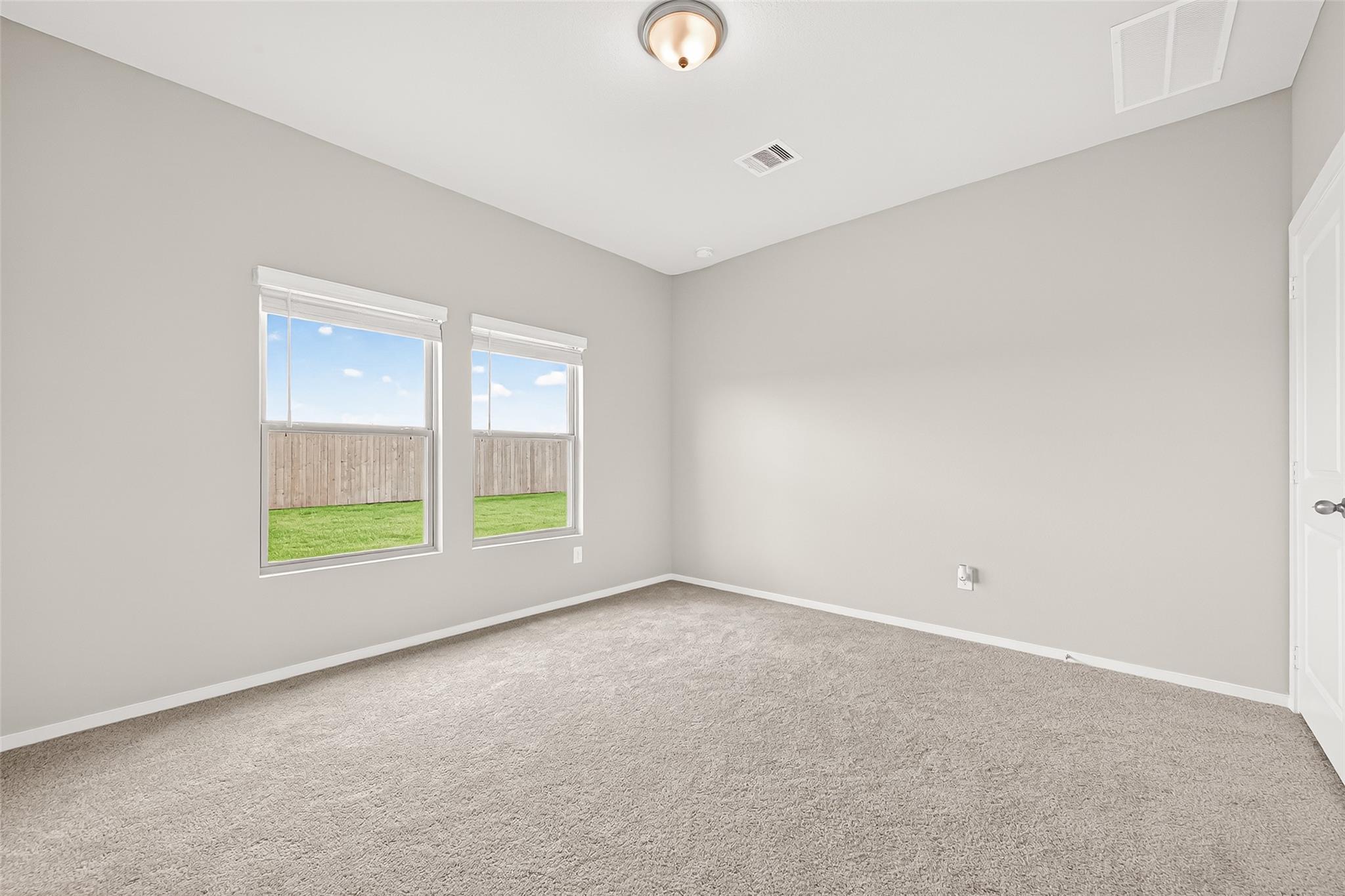 This is a bright, carpeted room with neutral walls, featuring two windows that provide natural light and a view of a fenced backyard.