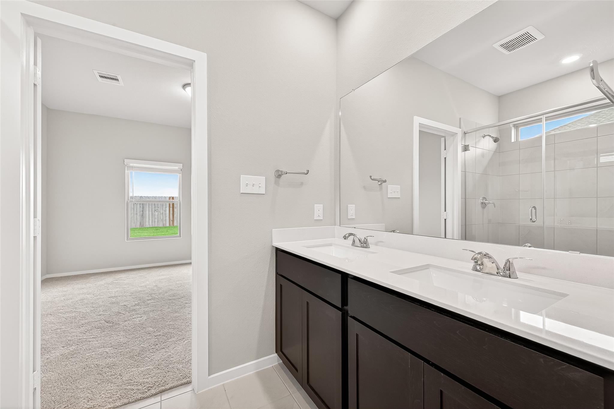This photo showcases a modern bathroom with a double vanity featuring sleek faucets and ample storage. It leads to a carpeted bedroom with a window providing natural light and a view of the fenced yard.