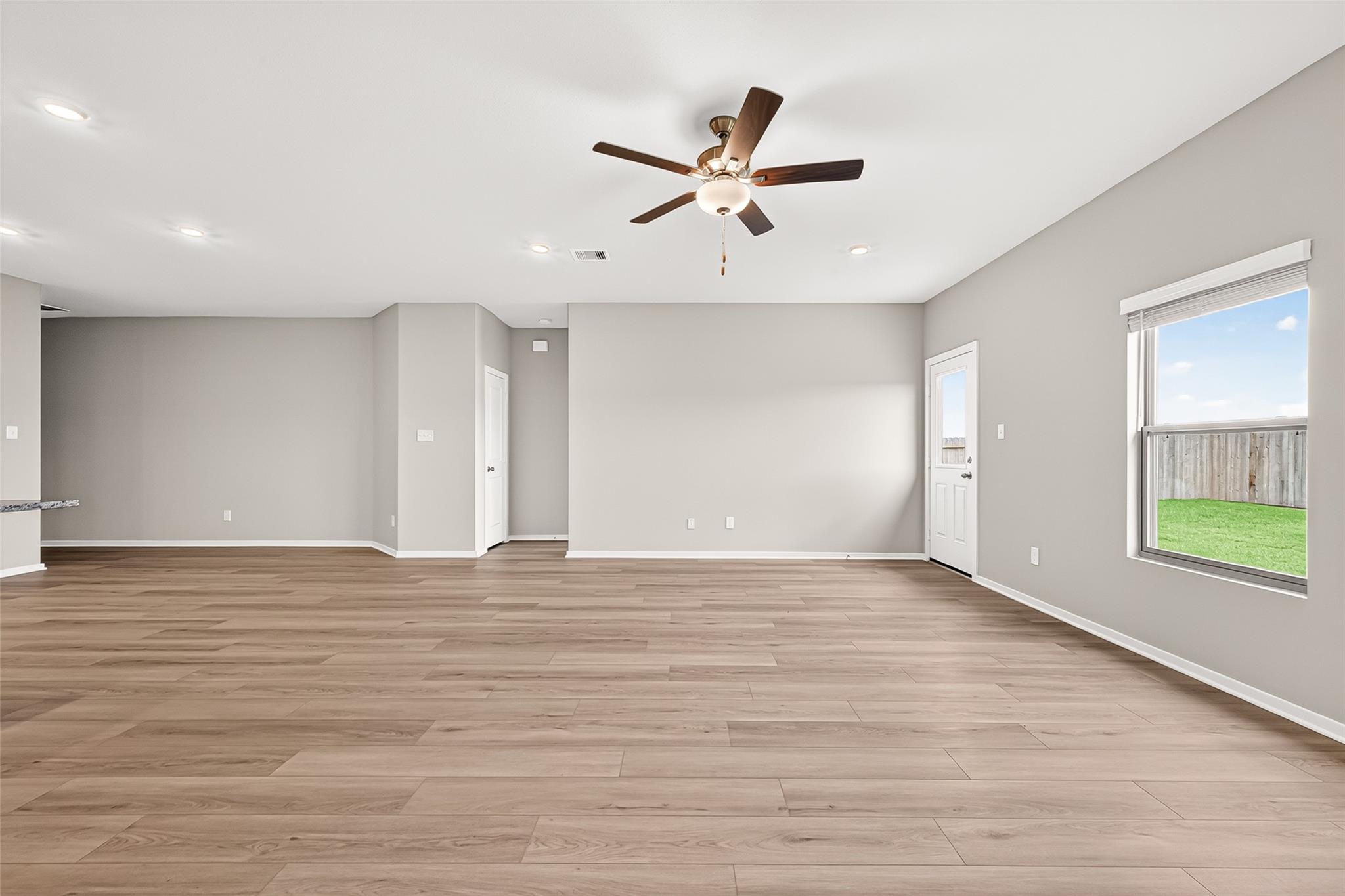 This spacious living area features light wood flooring, neutral gray walls, recessed lighting, and a ceiling fan. A large window and door provide ample natural light and access to a fenced backyard.