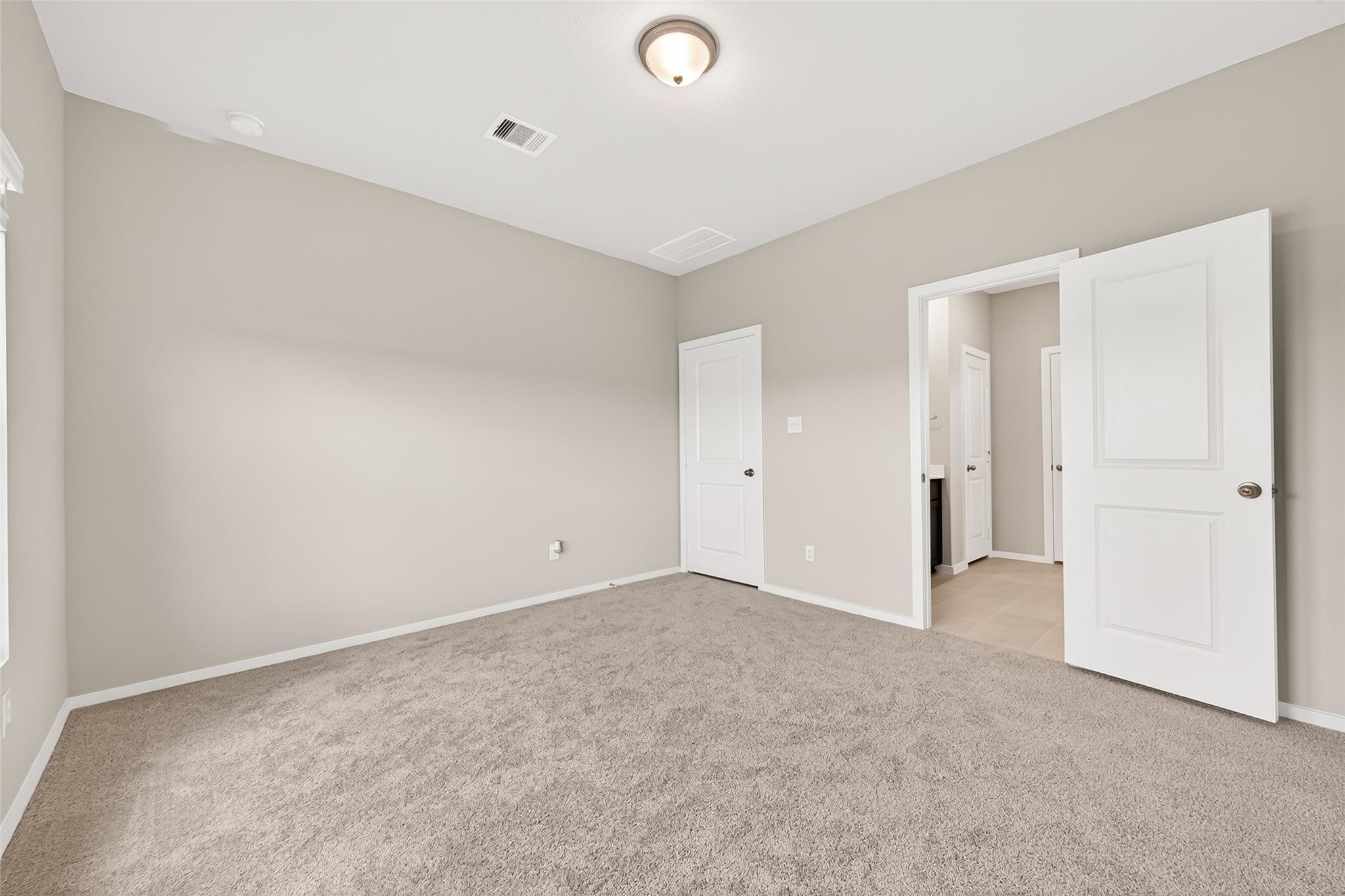This is a spacious, carpeted bedroom with neutral walls, a ceiling light, and white doors leading to a bathroom and closet.