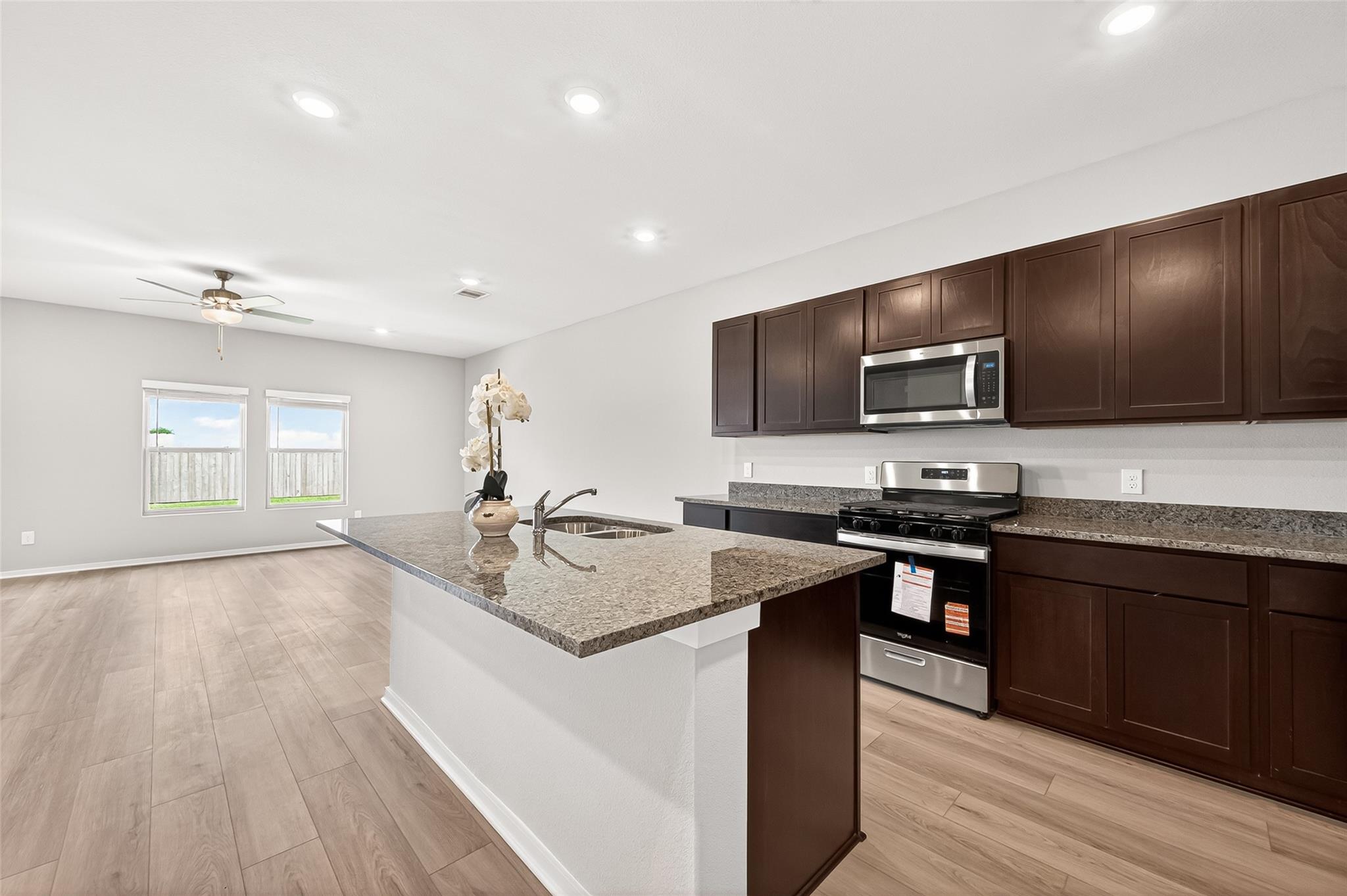This open-concept kitchen and living area features modern finishes with dark wood cabinets, granite countertops, and stainless steel appliances. The space is bright and airy, enhanced by recessed lighting, a ceiling fan, and large windows overlooking a fenced backyard.
