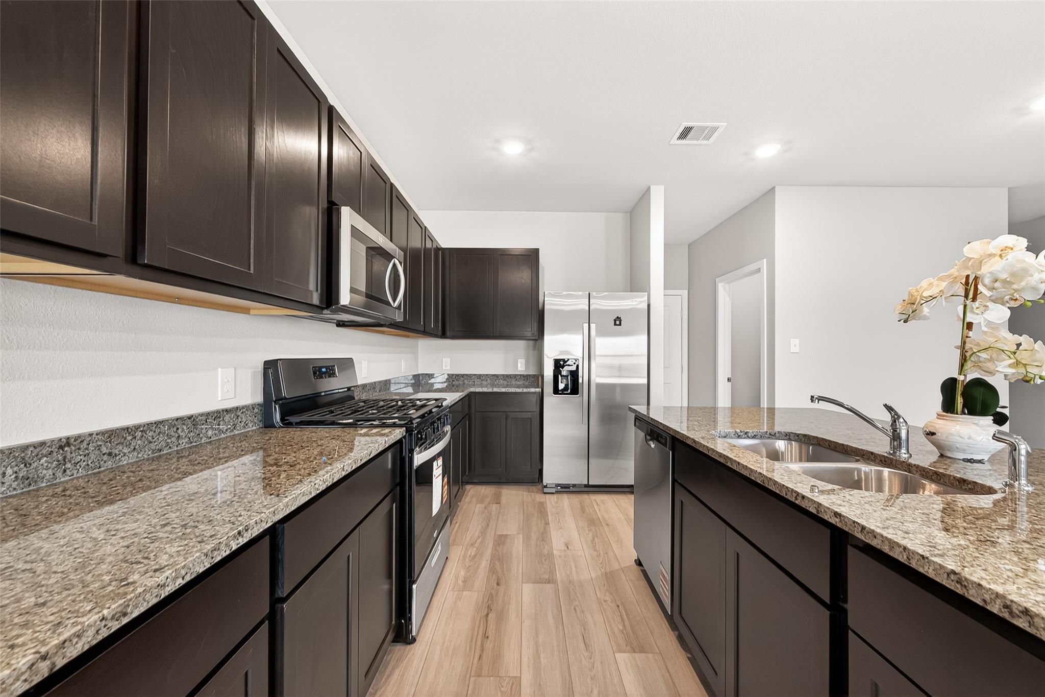This modern kitchen features dark wood cabinetry, granite countertops, and stainless steel appliances, including a fridge and oven. It has a spacious layout with a central island and double sink, complemented by light wood flooring and bright lighting.