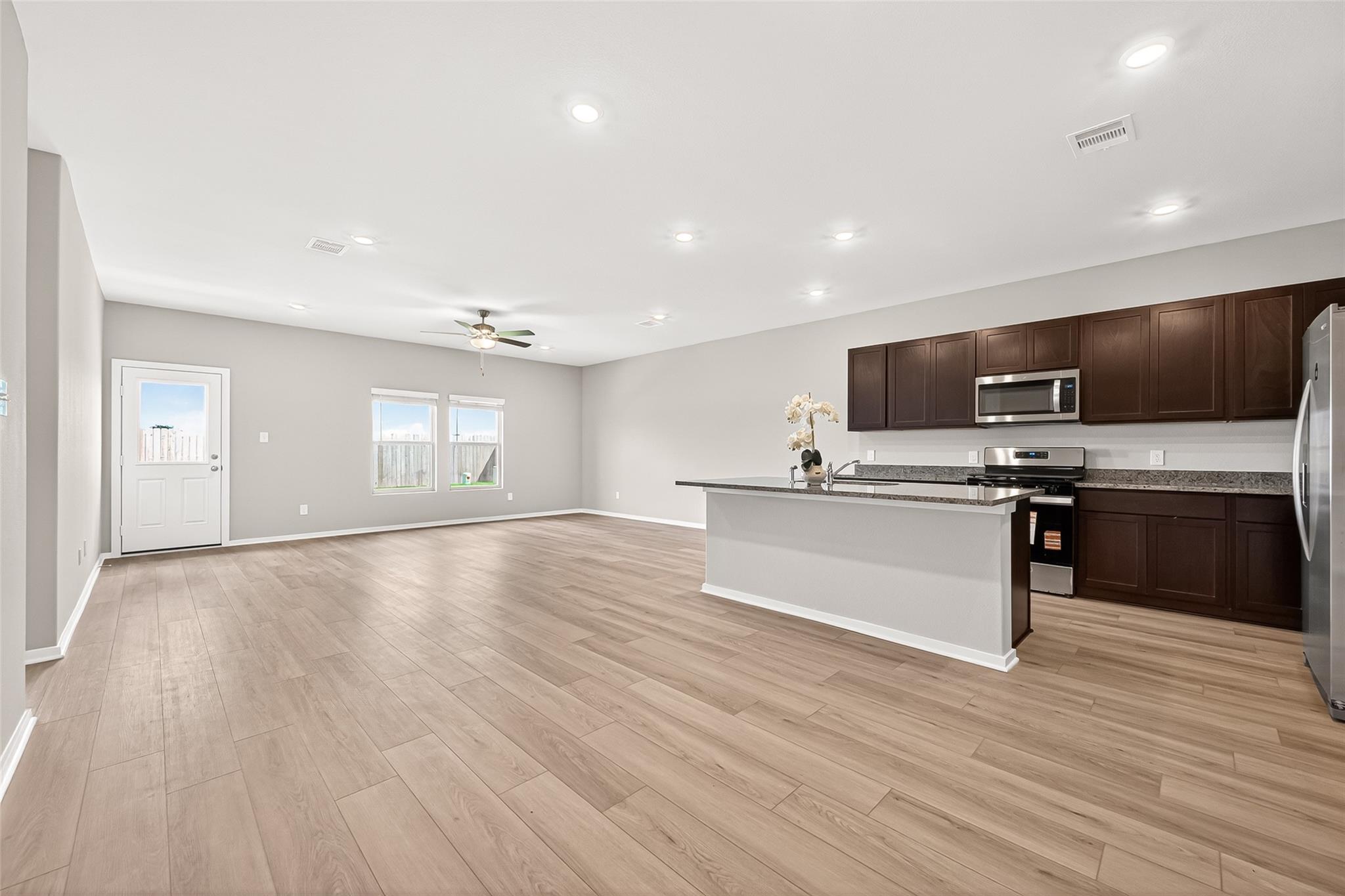 This spacious open-concept living area features modern wood flooring, ample natural light, and a sleek kitchen with dark cabinets, granite countertops, and stainless steel appliances. Ideal for entertaining and everyday living.