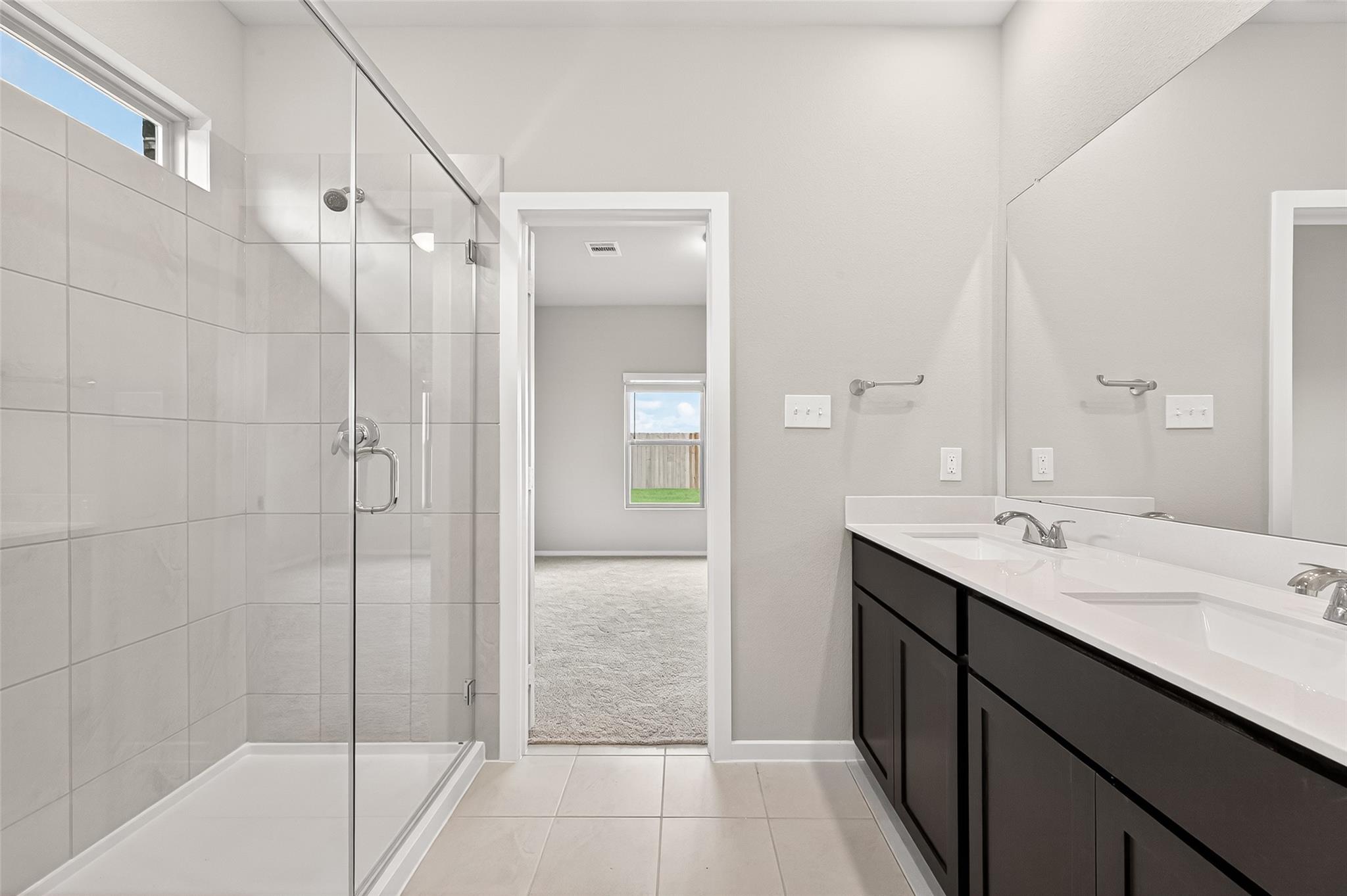 Modern bathroom featuring a spacious glass-enclosed shower and a double-sink vanity with sleek dark cabinetry. The room is brightly lit, with a neutral color palette, and opens to a carpeted area with a window view.