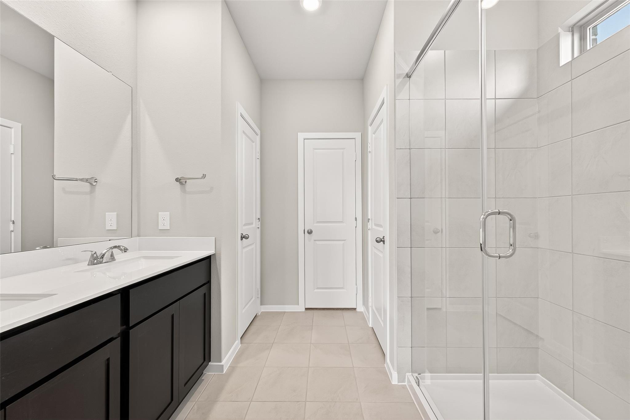 This modern bathroom features a sleek double vanity with dark cabinetry, a large mirror, and a spacious glass-enclosed shower. Bright tiles and neutral tones create an inviting and clean atmosphere.