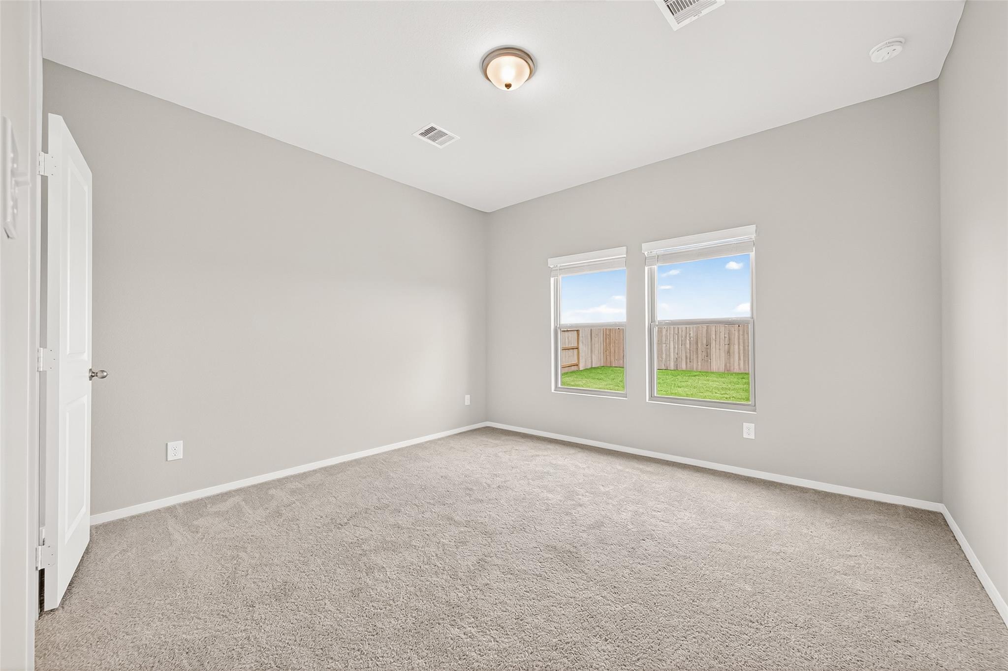 This room is bright and airy, featuring neutral walls and carpet, a ceiling light, and two windows offering a view of a fenced backyard.