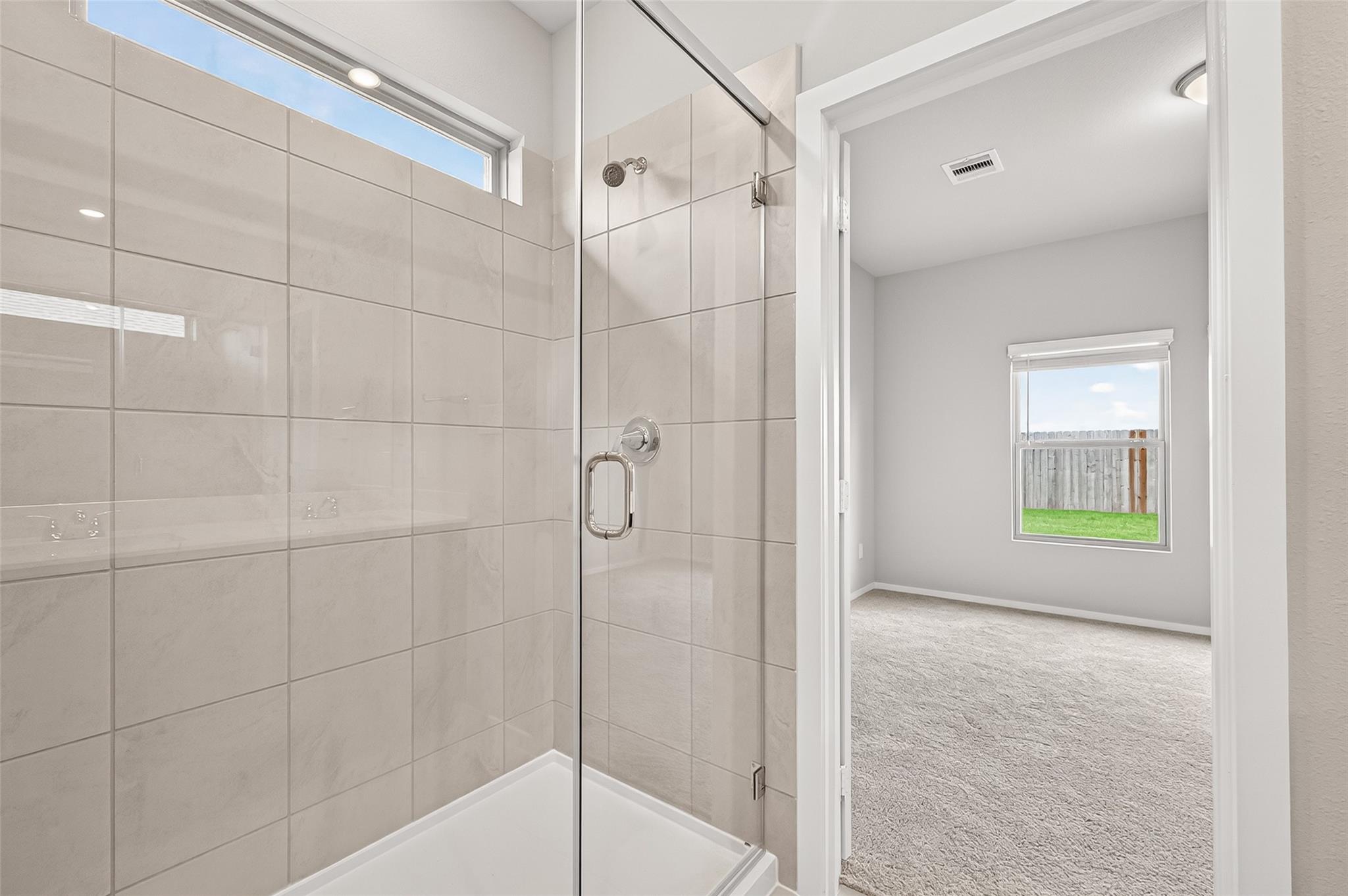 This photo shows a modern bathroom with a glass-enclosed shower and light beige tile. Adjacent is a carpeted room with a window overlooking a fenced yard, offering natural light and privacy.