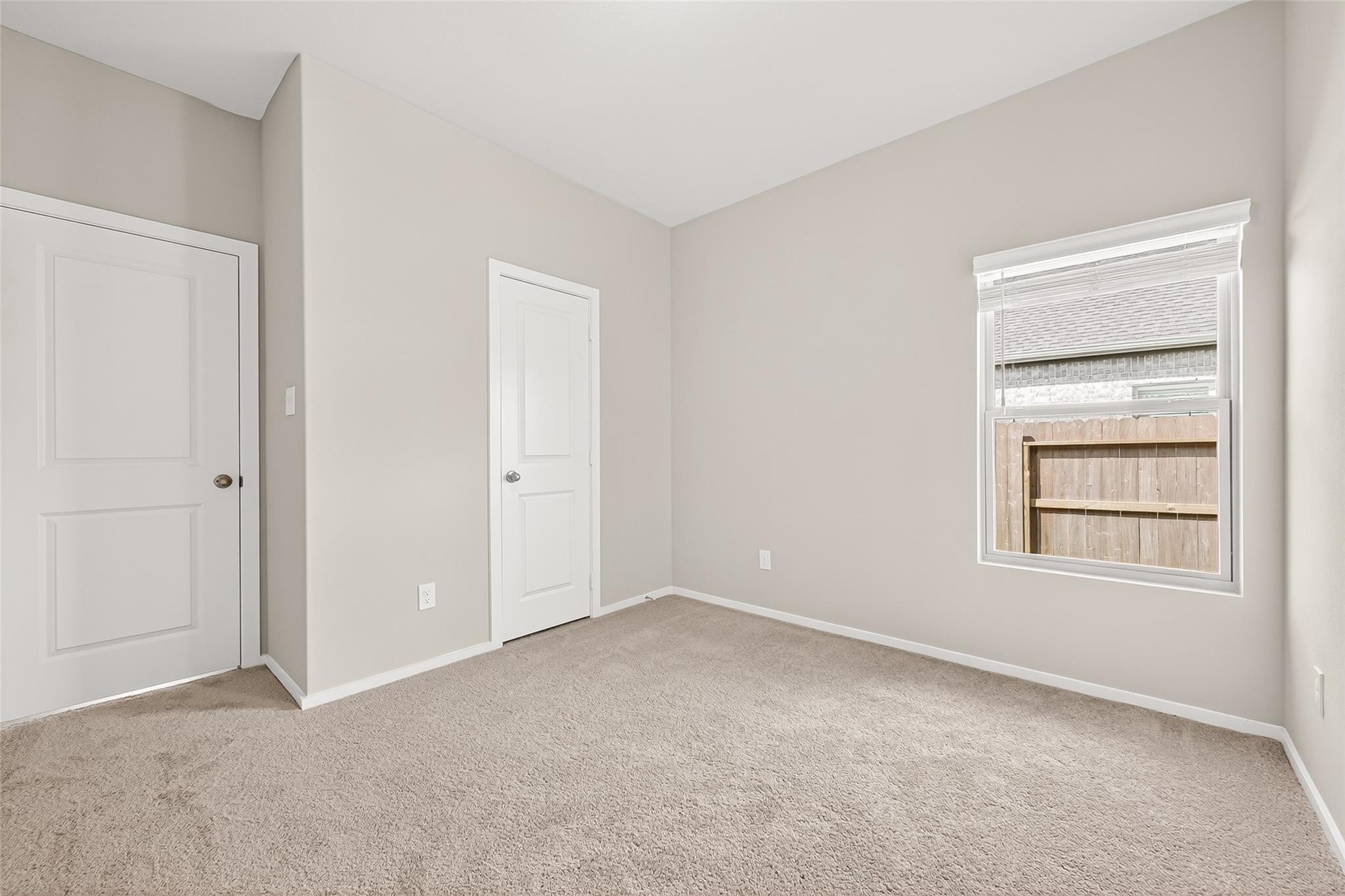 This is a bright, neutral-toned room with carpeted flooring, featuring a window with blinds and two white doors. Ideal for a bedroom or home office.