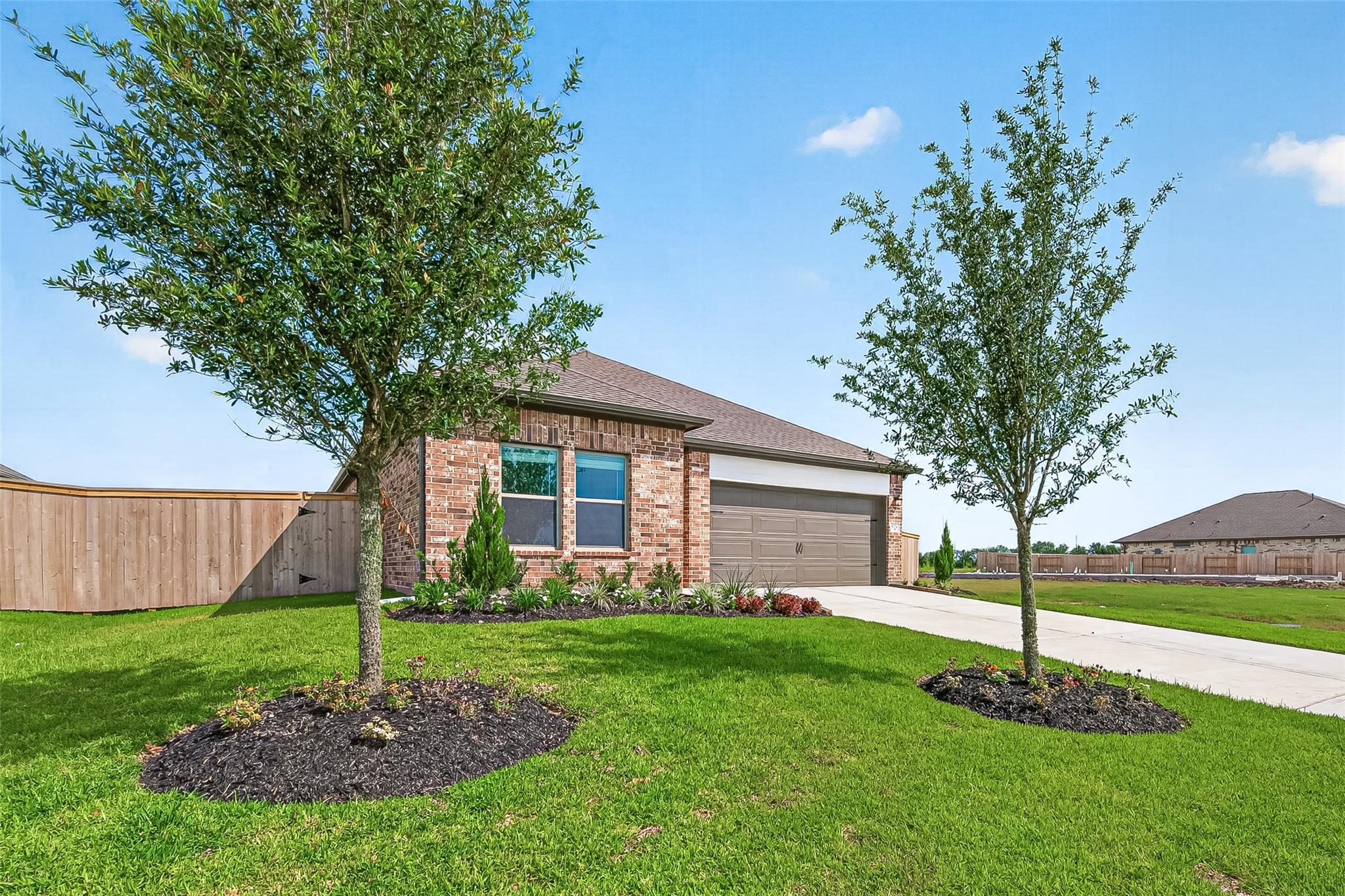 Charming brick home with a two-car garage, nestled in a well-maintained yard featuring young trees and landscaped flower beds. The property is enclosed by a wooden fence for added privacy.