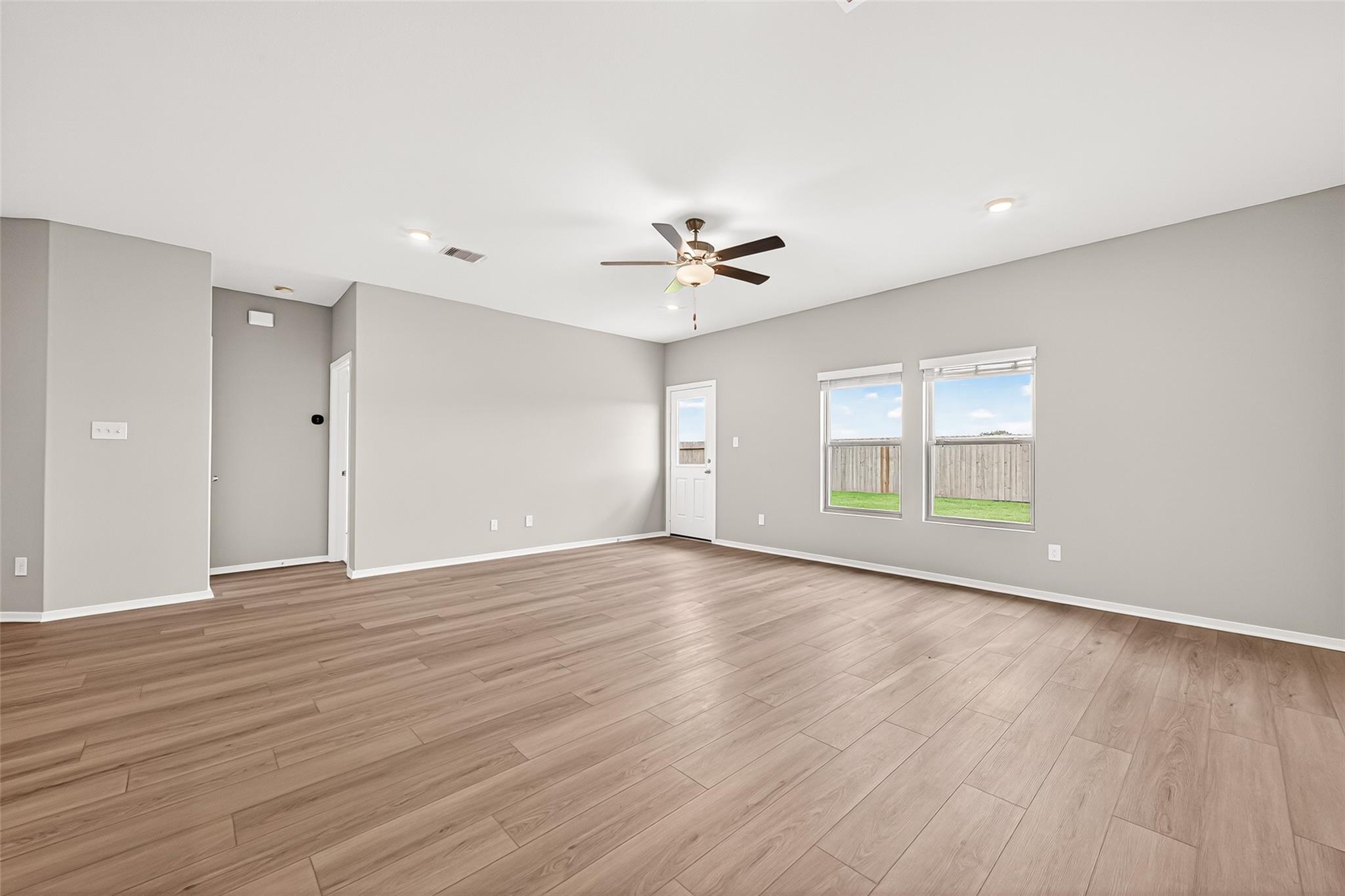 This spacious, light-filled room features modern wood flooring, neutral gray walls, and a ceiling fan. Large windows offer views of the backyard, and a door provides easy outdoor access.