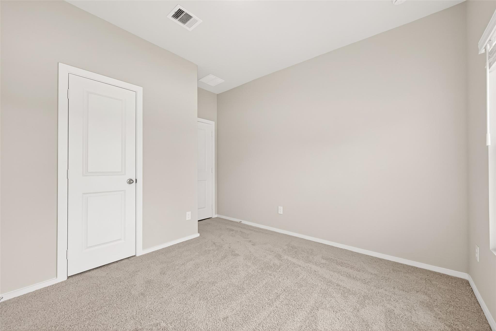 This is a bright, neutral-toned room with fresh carpet and a simple, modern aesthetic. It features two white doors, presumably for a closet and entry, and a small window allowing natural light to enter. Ideal for use as a bedroom or a home office.