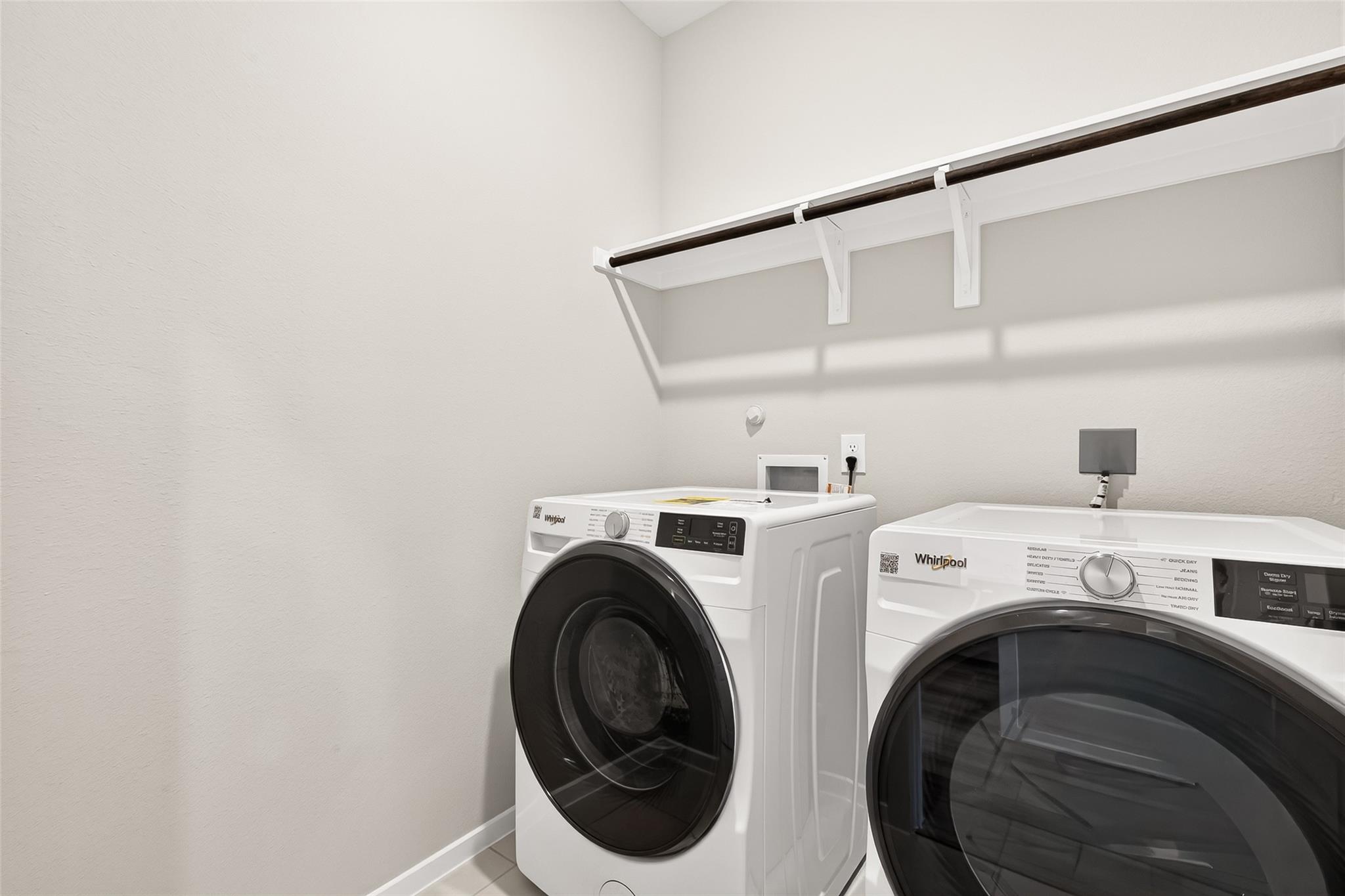 This photo shows a compact laundry room with modern appliances, featuring a front-loading washer and dryer. There's a wall-mounted shelf above for additional storage, with clean, neutral walls.