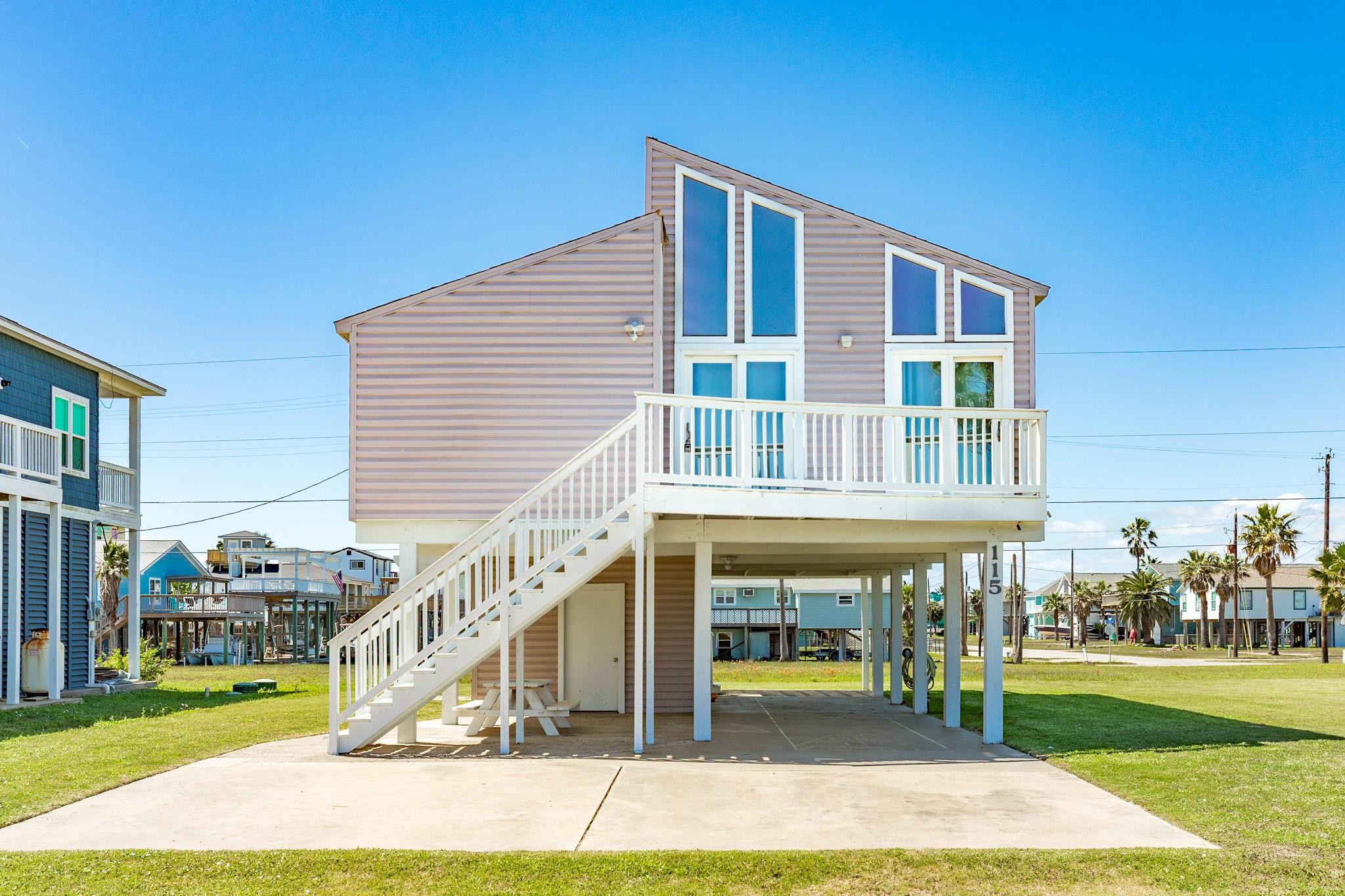 Front Exterior – Elevated coastal home featuring a distinctive sloped roofline and expansive windows that flood the interior with natural light. A spacious upper deck provides the perfect spot for outdoor seating and enjoying the coastal breeze.